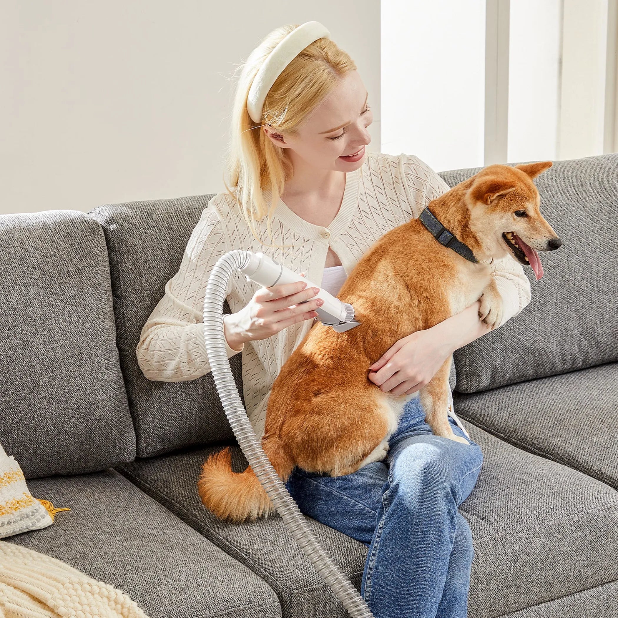 Woman sitting on a couch with a dog, using a hair dryer.
